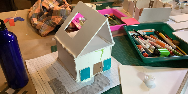 A model is presented with a square base and slanted triangle roof on a drafting table in Wright’s studio