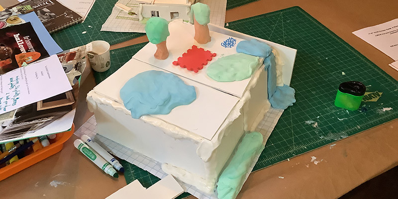 On one of Wright’s drafting tables, a camper has created a square shaped model home with handmade trees, waterfalls, and water features made of colored model magic on the roof