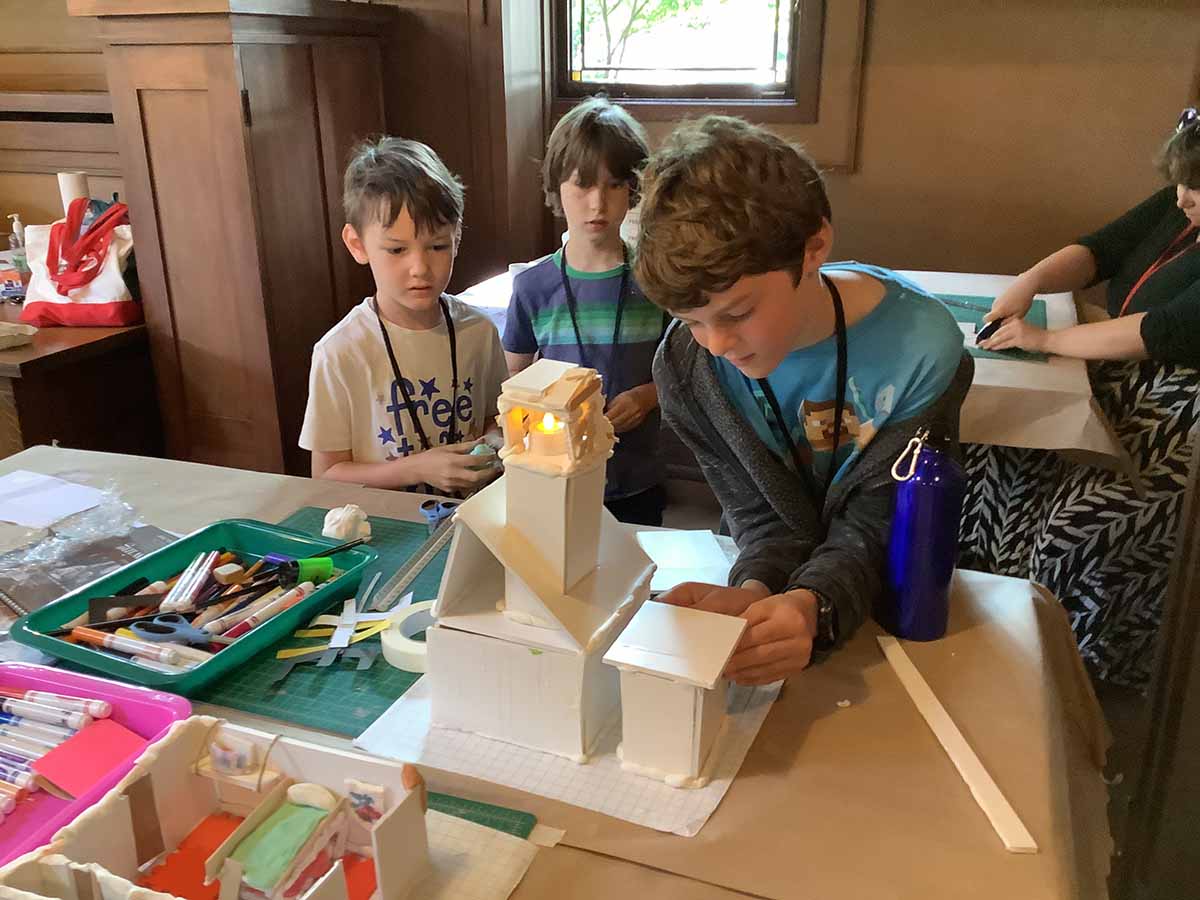 Two young campers look carefully at an older camper’s model as he puts on the finishing touches